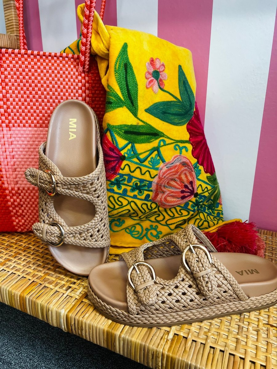 Pair of brown sandals with 'MIA' branding next to a colorful bag on a striped background