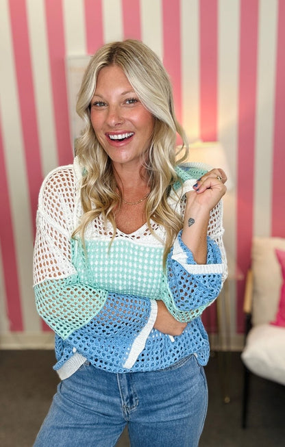 A smiling blonde woman wearing a colorful crochet knit top with white, mint green, and light blue sections, paired with blue jeans, standing in front of a pink and white striped wall.