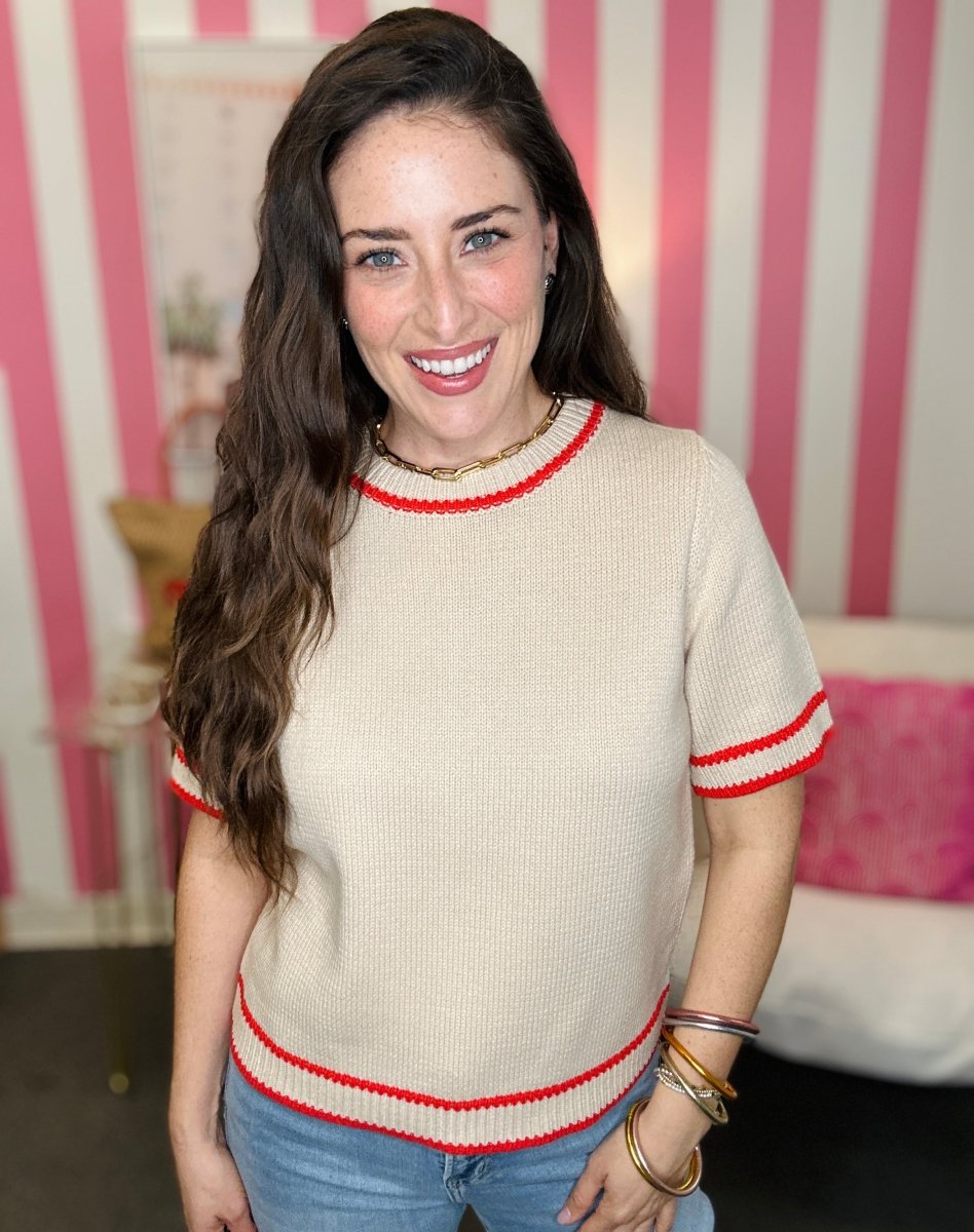 Woman wearing a beige sweater with red trim in a room with pink and white striped wallpaper.