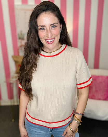 Woman wearing a beige sweater with red trim in a room with pink and white striped wallpaper.
