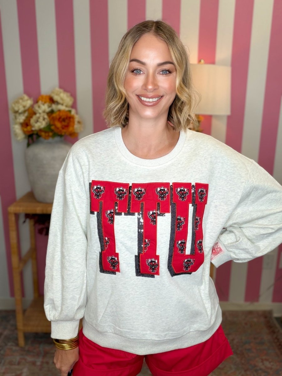 A smiling woman wearing a light gray sweatshirt with a large red and black 'ITD' graphic on the front, paired with bright red pants, standing in front of a pink and white striped wall.