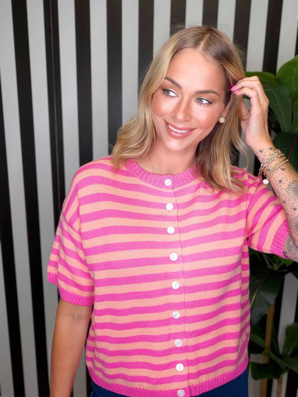 A woman wearing a pink and peach striped short-sleeve knit cardigan sweater with white buttons, posing in front of a black and white striped wall.