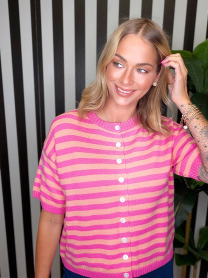 A woman wearing a pink and peach striped short-sleeve knit cardigan sweater with white buttons, posing in front of a black and white striped wall.