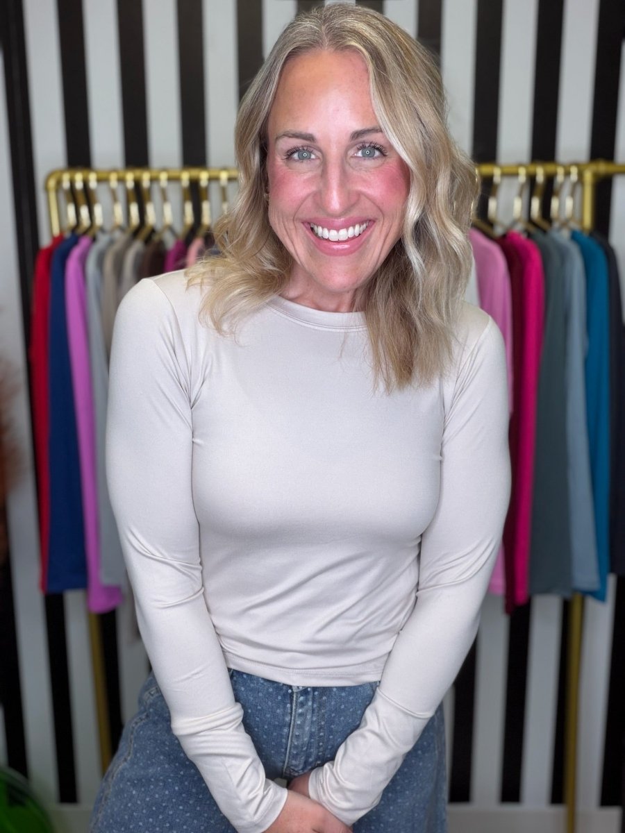 A smiling blonde woman wearing a fitted, light beige long-sleeve top and blue jeans, standing in front of a clothing rack filled with colorful garments against a black and white striped wall.