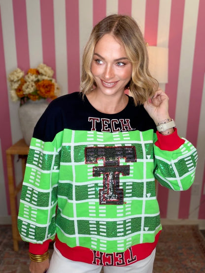 A woman wearing a festive Texas Tech themed sweater with a black upper section, bright green and white football field patterned sleeves and body, and a large sequined 'T' on the chest, posed against a pink and white striped background.