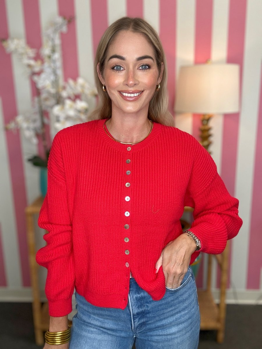 A woman wearing a bright red waffle-knit button-front cardigan sweater and blue jeans, standing in front of a pink and white striped wall with a lamp and decorative flowers in the background.