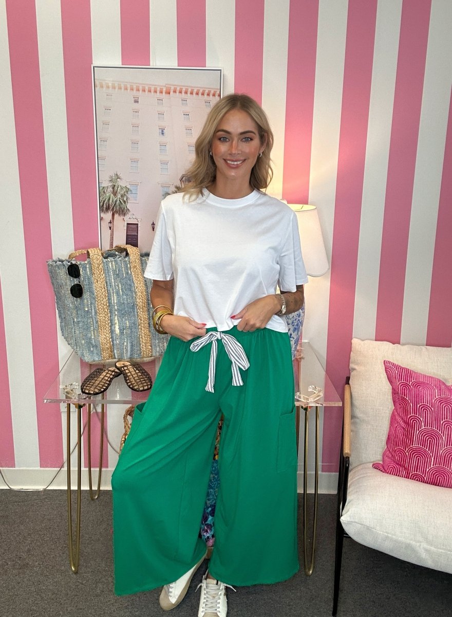 A woman stands in a room with pink and white striped wallpaper, wearing a white t-shirt and bright green wide-leg pants with a white striped drawstring tie. She is posing with a denim and gold striped tote bag on a glass side table.