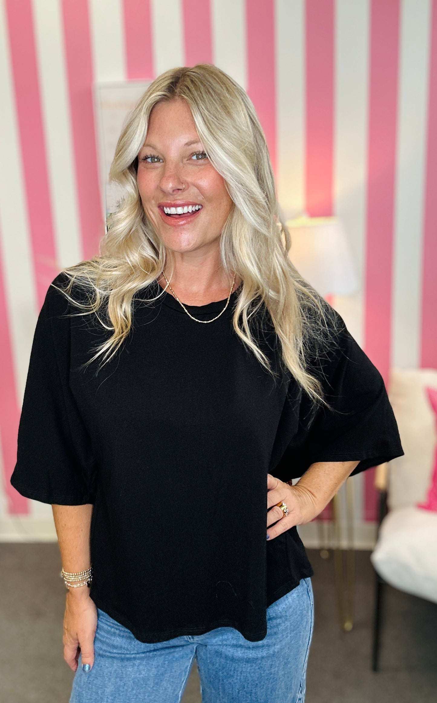A woman with long blonde hair wearing a black short-sleeve top and blue jeans, standing in front of a pink and white striped wall.
