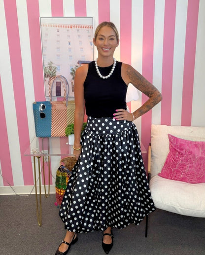 A woman stands in a room with pink and white striped walls, wearing a sleeveless black top and a black midi skirt with white polka dots. She is accessorized with a pearl necklace and black pointed flats.