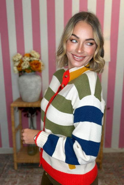 A woman wearing a colorful striped cardigan sweater with green, white, blue, and red horizontal stripes, standing in front of a pink and white striped wall.