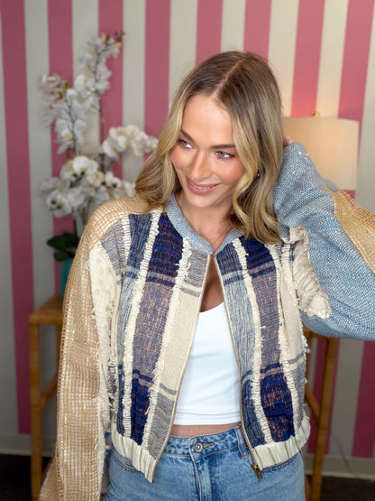 A woman wearing a textured patchwork knit jacket with beige, blue, and navy plaid panels, styled with a white tank top and blue jeans, standing in front of a pink and white striped wall.