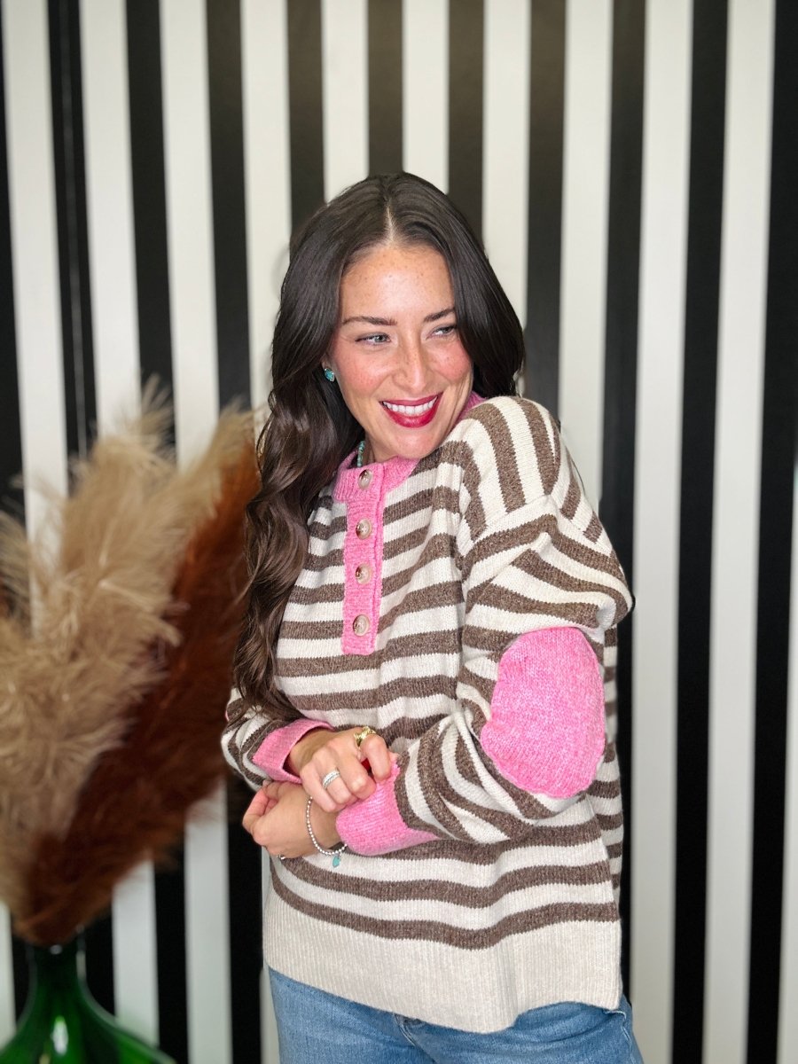 A woman smiling and wearing a brown and beige striped knit sweater with bright pink elbow patches and a pink button placket, paired with blue jeans, set against a black and white striped background.
