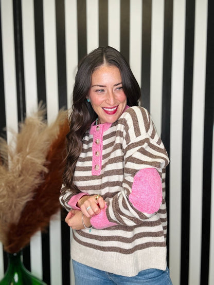 A woman smiling and wearing a brown and beige striped knit sweater with bright pink elbow patches and a pink button placket, paired with blue jeans, set against a black and white striped background.