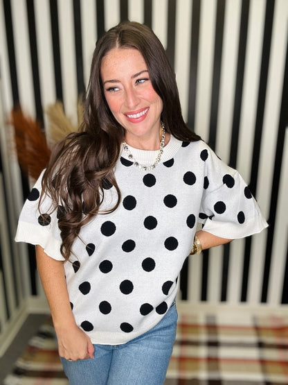 A woman wearing a white short-sleeve knit top with black polka dots, paired with blue jeans, standing in front of a black and white striped background.