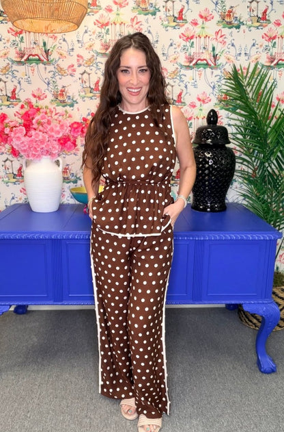 A woman modeling a brown sleeveless jumpsuit with white polka dots and white side stripes, standing in front of a blue cabinet and floral wallpaper.