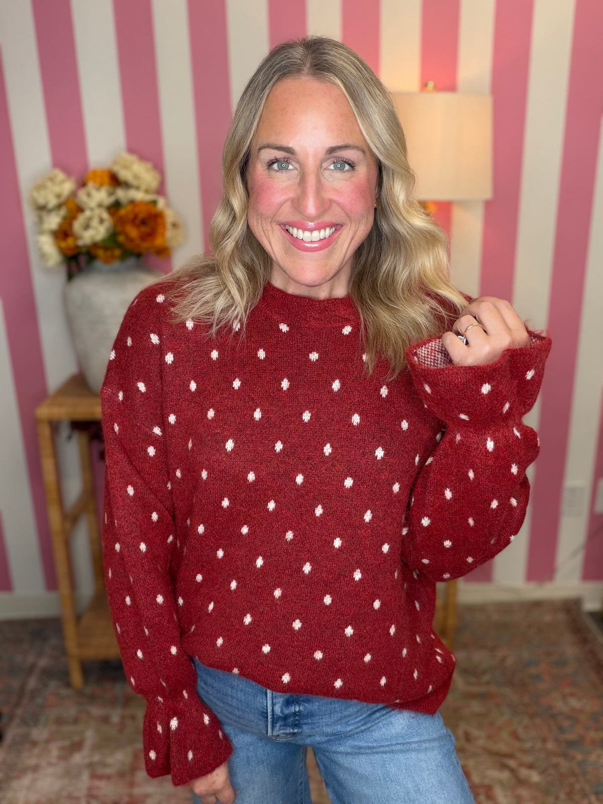 A smiling woman with blonde hair wearing a red knit sweater with a white dotted pattern and blue jeans, standing in front of a pink and white striped wall.