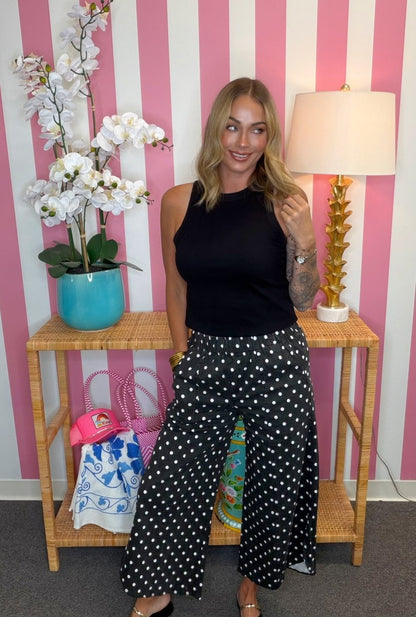 A woman stands indoors wearing a sleeveless black top and black wide-leg pants with white polka dots. She is posed in front of a pink-and-white striped wall, with a wooden side table holding a turquoise orchid pot, a pink handbag, and a lamp on a white base.