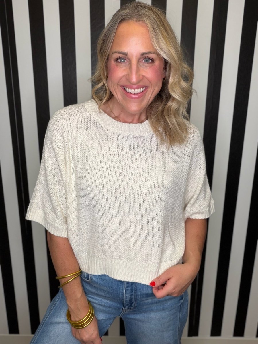 A smiling blonde woman wearing a cream-colored short-sleeve knit sweater and blue jeans, standing in front of a black and white vertically striped wall.
