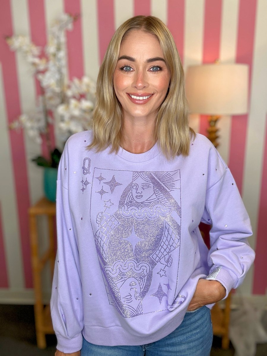 A smiling blonde woman wearing a lavender crewneck sweatshirt with a large silver-toned graphic of a playing card on the front, paired with blue jeans, standing in front of a pink and white striped wall.