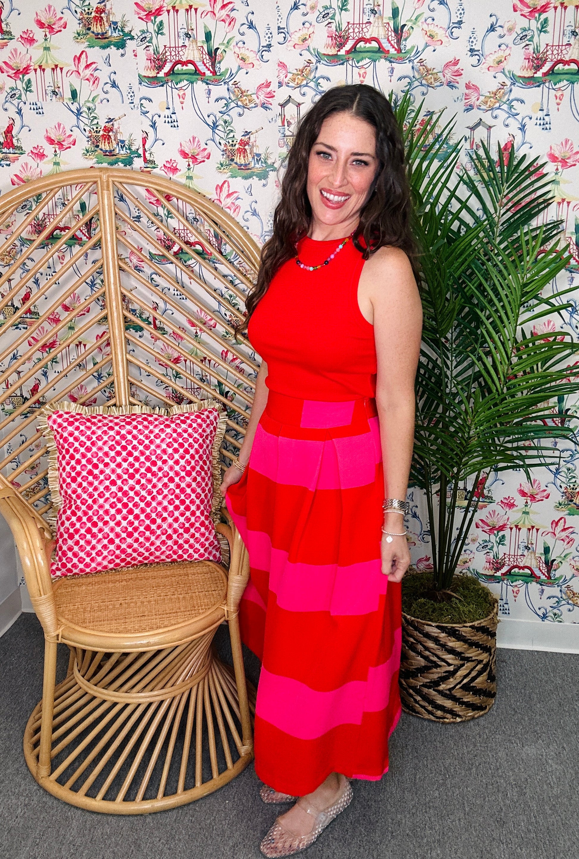 Woman in a red and pink dress standing next to a wicker chair with a pink cushion and a plant against a patterned wall.