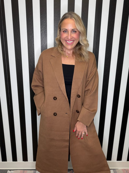 Woman in a brown trench coat standing in front of a black and white striped wall smiling.