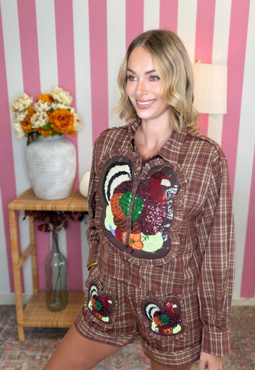 A woman wearing a brown plaid long-sleeve shirt and matching shorts set, both decorated with large sequined turkeys on the front, posed in front of a pink and white striped wall.