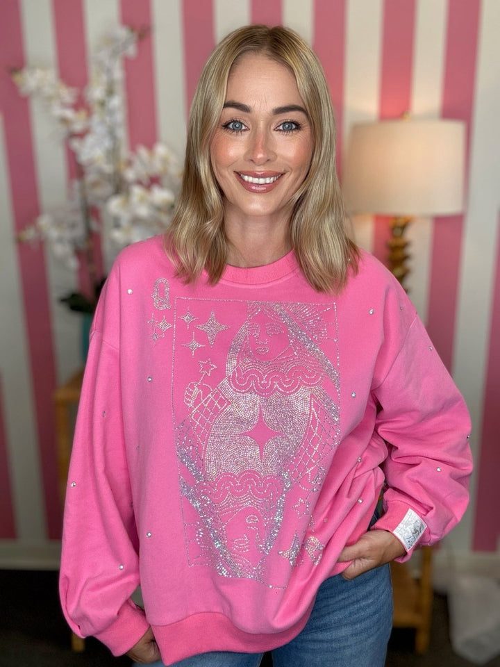 A smiling blonde woman wearing an oversized bright pink sweatshirt with a large silver glitter graphic on the front, posed in front of a pink and white striped wall.