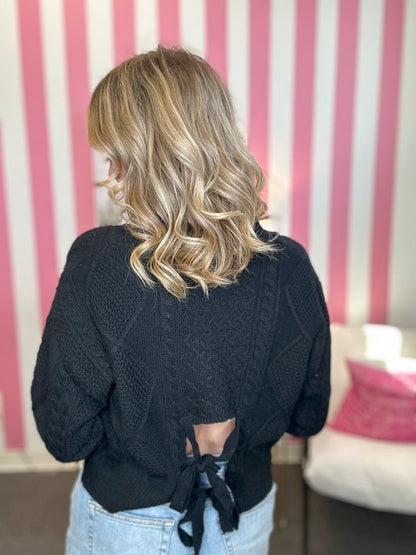 A woman viewed from behind wearing a black cable-knit sweater with a tie-back detail, paired with light blue jeans, set against a pink and white striped wall.