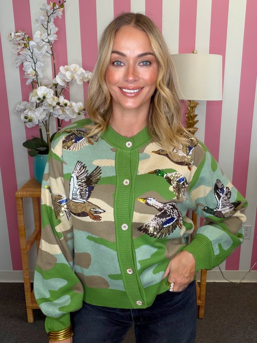 A woman wearing a green camouflage knit cardigan with embroidered ducks, posed in front of a pink and white striped wall.