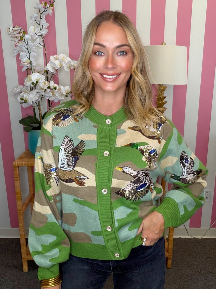 A woman wearing a green camouflage knit cardigan with embroidered ducks, posed in front of a pink and white striped wall.