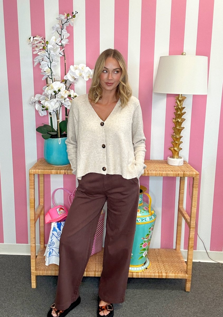 A woman stands in front of a pink and white striped wall, wearing a cream knit button-front cardigan and brown wide-leg pants. She is posed in front of a wooden side table holding a turquoise orchid pot and a decorative lamp.