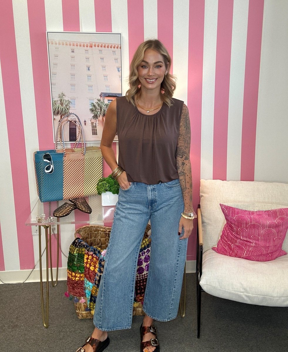 A woman stands indoors wearing a sleeveless brown top and wide-leg light blue jeans, with a pink-and-white striped wall behind her and a white chair with a pink pillow to the right.