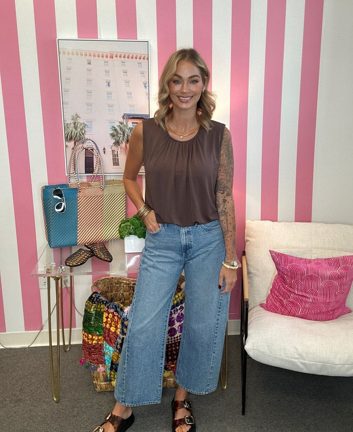 A woman stands indoors wearing a sleeveless brown top and wide-leg light blue jeans, with a pink-and-white striped wall behind her and a white chair with a pink pillow to the right.