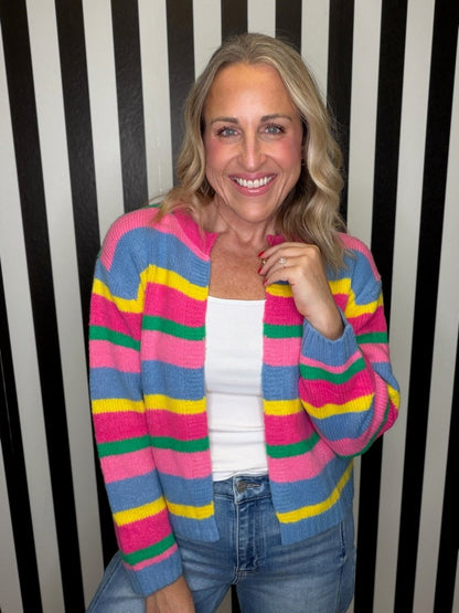 A smiling woman with blonde hair wearing a colorful striped knit cardigan over a white tank top and blue jeans, standing in front of a black and white vertically striped wall.