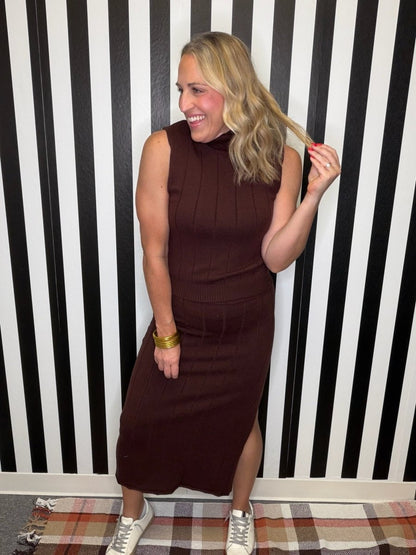 A woman modeling a sleeveless, ribbed brown mock-neck midi dress with a side slit, standing in front of a black and white striped wall.