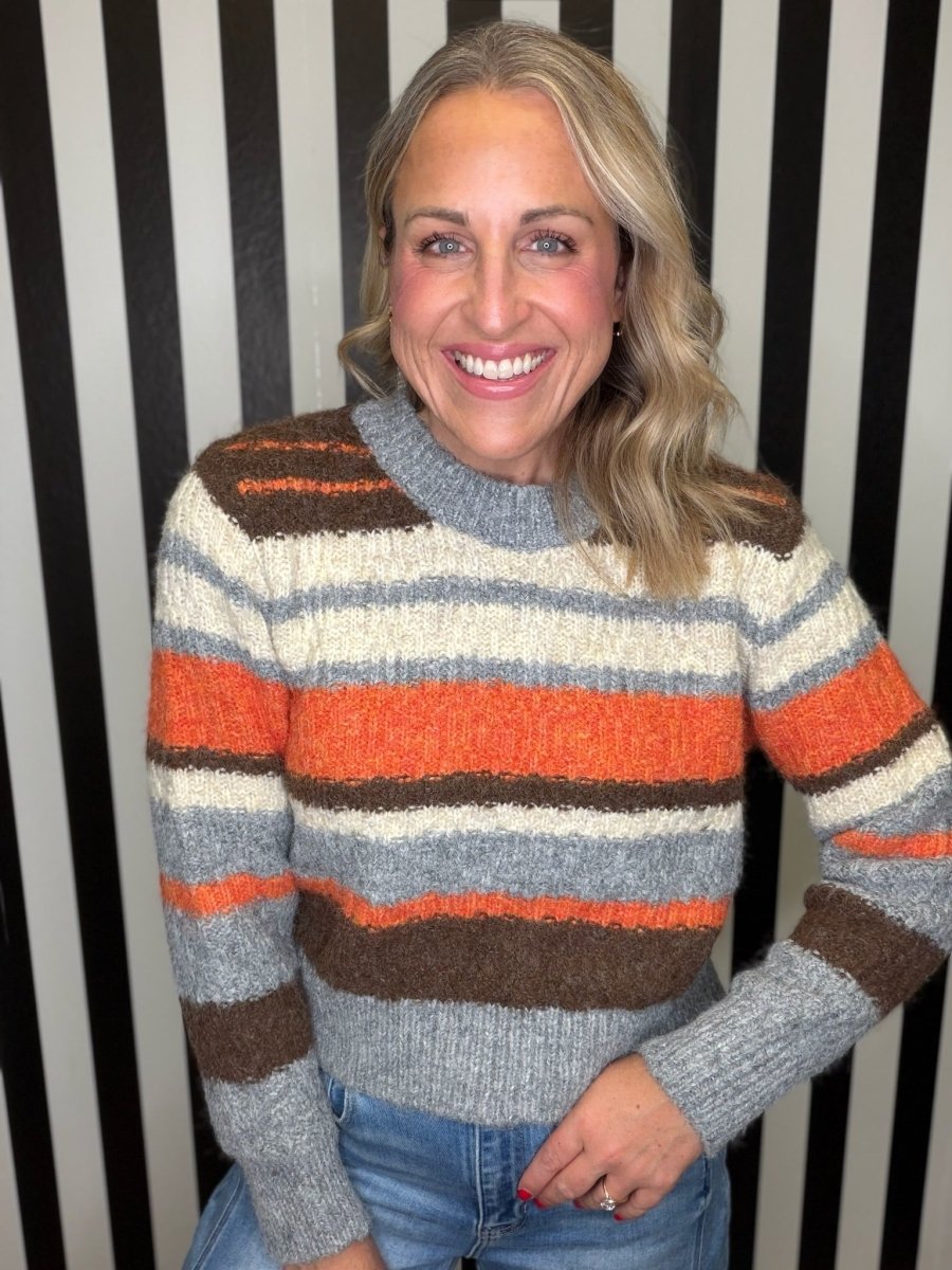 A smiling woman wearing a chunky knit striped sweater with horizontal bands of gray, cream, orange, and brown, paired with blue jeans, standing against a black and white vertically striped background.