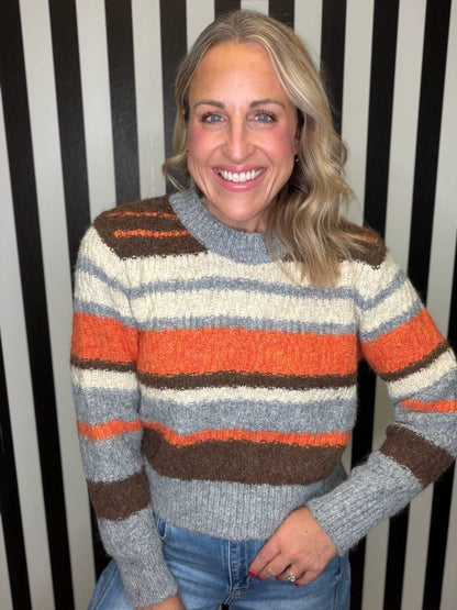 A smiling woman wearing a chunky knit striped sweater with horizontal bands of gray, cream, orange, and brown, paired with blue jeans, standing against a black and white vertically striped background.