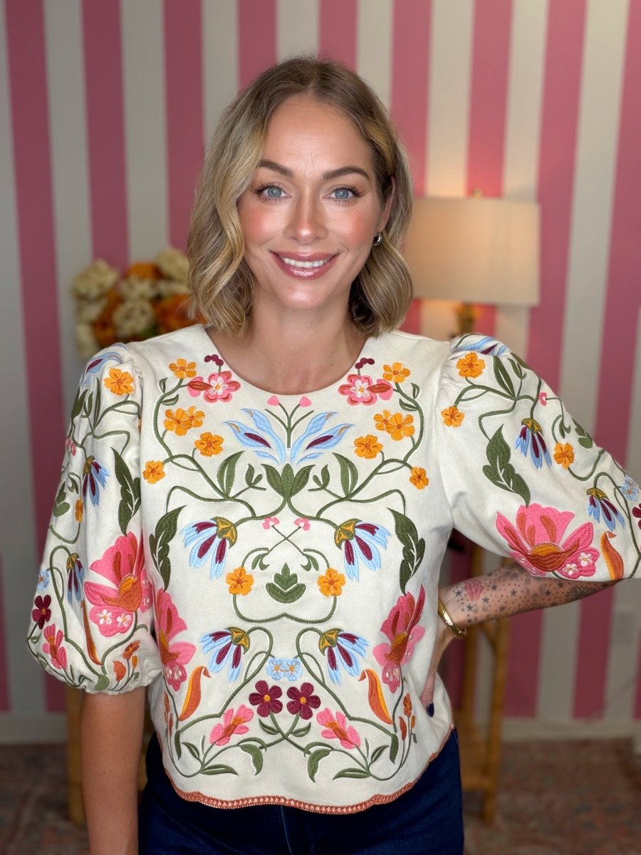 A woman wearing a cream-colored blouse with vibrant, multicolored floral embroidery and puff sleeves, standing in front of a pink and white striped wall.