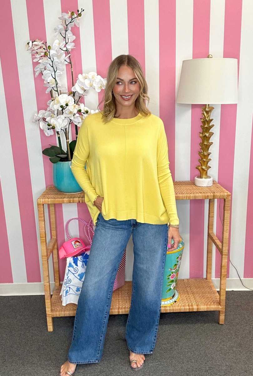 A woman stands in a room with pink and white striped wallpaper, wearing a bright yellow long-sleeve top and blue wide-leg jeans. She is posed in front of a wooden side table with a lamp, a vase of white flowers, and colorful bags.