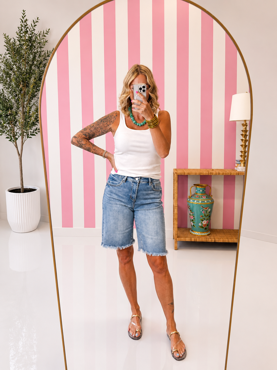 Woman taking a selfie in front of a pink and white striped wall with a plant and shelf in the background.
