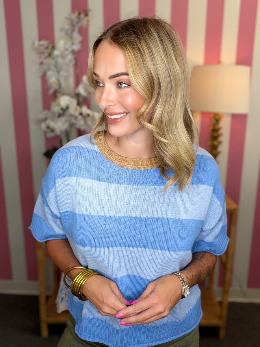 A woman with blonde hair wearing a light blue and white striped knit sweater with a beige crew neck, standing in front of a pink and white striped wall.