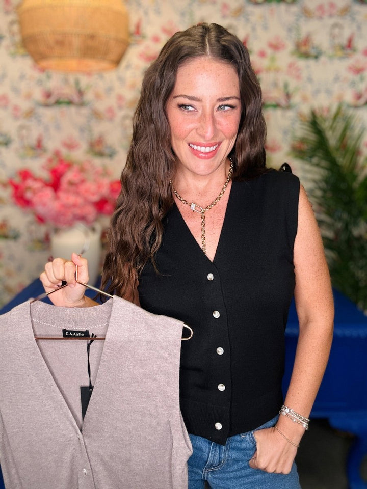 A woman with long brown hair smiling while holding a light gray knit vest on a hanger. She is wearing a black sleeveless button-front top and blue jeans, standing in front of a floral wallpaper background.