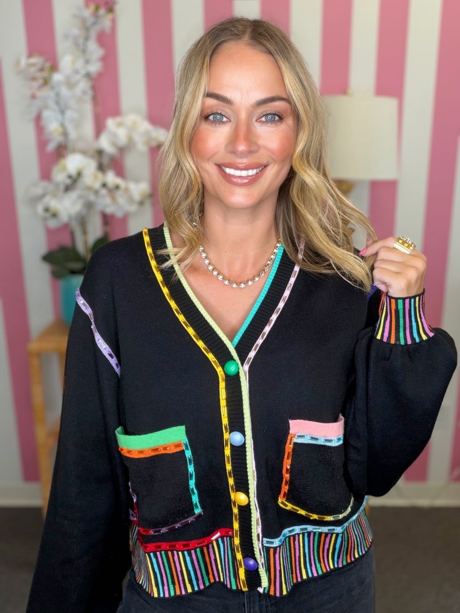 A smiling blonde woman wearing a black knit cardigan with vibrant multicolor striped and patchwork accents on the pockets, trim, and cuffs, posed against a pink and white striped wall.