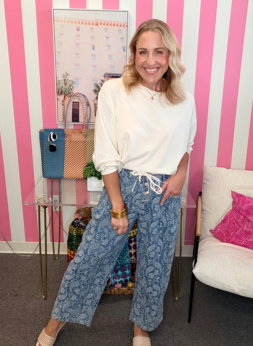 A woman posing in a white long-sleeve top and blue paisley wide-leg pants with a white drawstring, set against a pink and white striped wall.