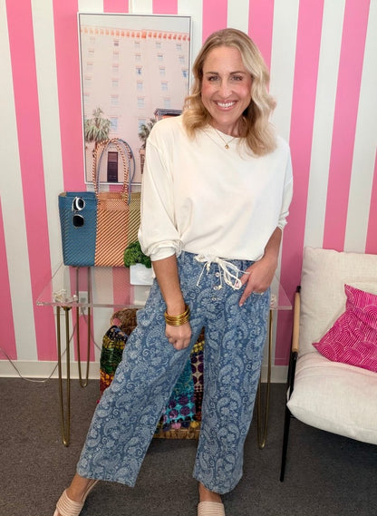 A woman posing in a white long-sleeve top and blue paisley wide-leg pants with a white drawstring, set against a pink and white striped wall.
