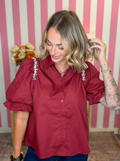 A woman wearing a burgundy short-sleeve button-up blouse with ornate silver embellishments on the shoulders, paired with dark blue jeans, standing in front of a pink and white striped wall.