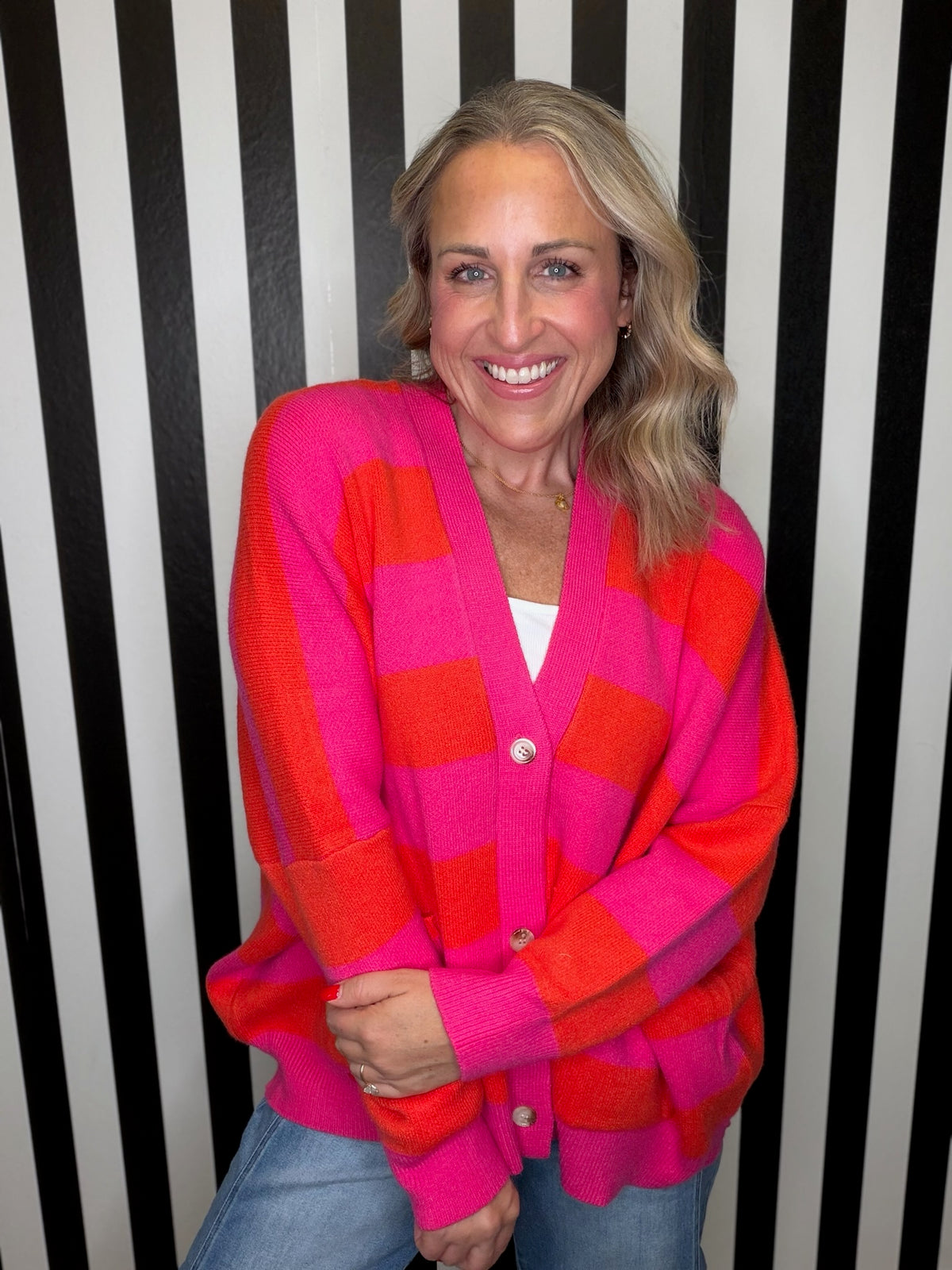 A smiling woman with blonde hair wearing a bright pink and orange checkered cardigan over a white top and blue jeans, standing in front of a black and white striped background.