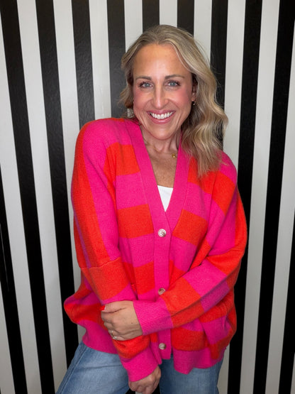 A smiling woman with blonde hair wearing a bright pink and orange checkered cardigan over a white top and blue jeans, standing in front of a black and white striped background.