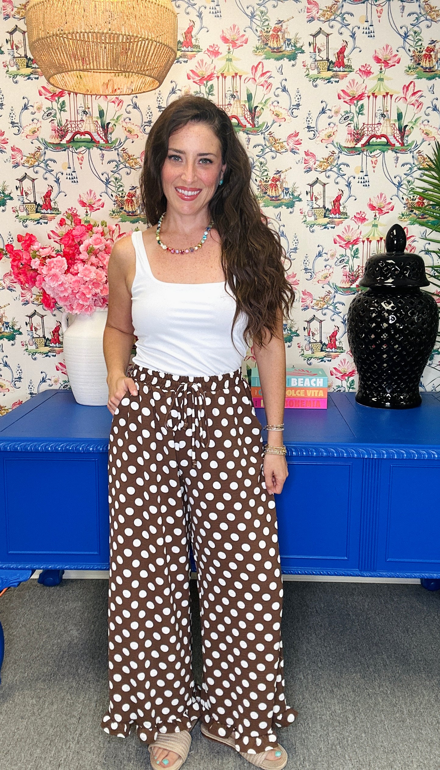 A woman modeling a white tank top and brown wide-leg pants with white polka dots, standing in front of a blue cabinet and floral wallpaper.