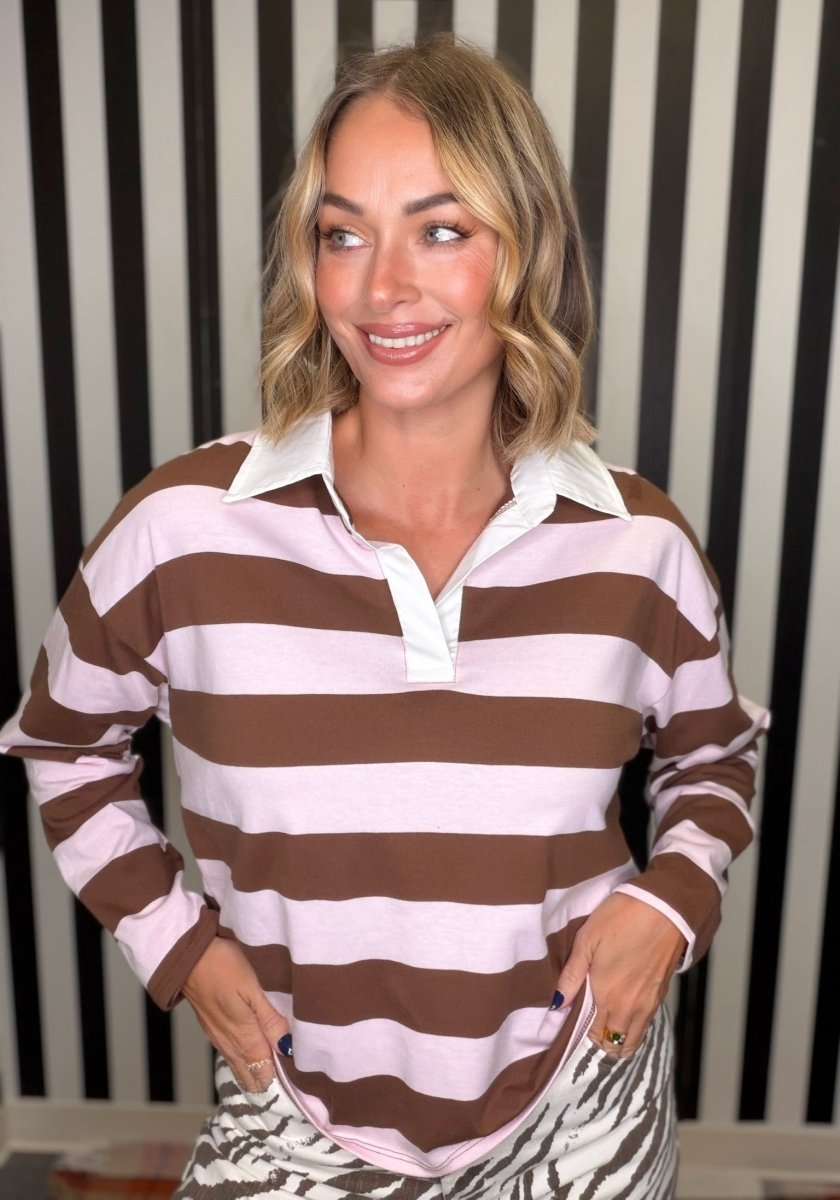 A woman wearing a brown and light pink horizontally striped long-sleeve polo shirt with a white collar, standing in front of a black and white striped wall.
